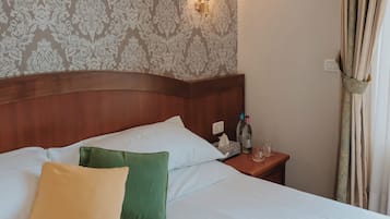 A neatly made bed with two pillows, a bedside table with a lamp and a bottle, and a patterned wallpaper.