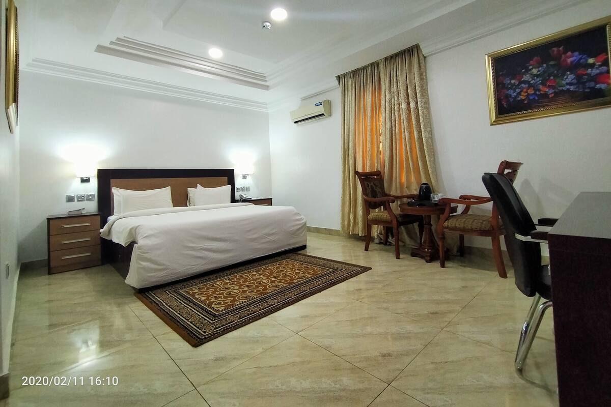 A hotel room with a bed, bedside tables, a desk with a chair, a painting, and a window with curtains.