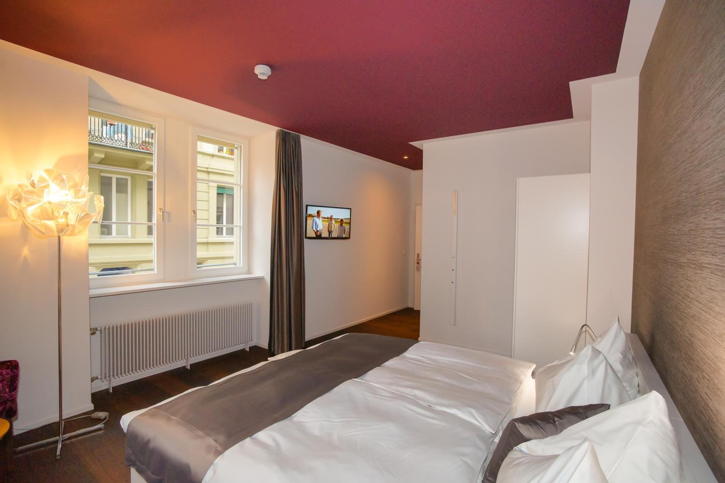 A hotel room with a bed, a radiator, a window with curtains, a television on the wall, and a ceiling light.