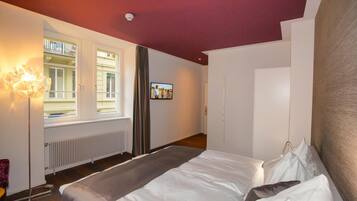 A hotel room with a bed, a radiator, a window with curtains, a television on the wall, and a ceiling light.
