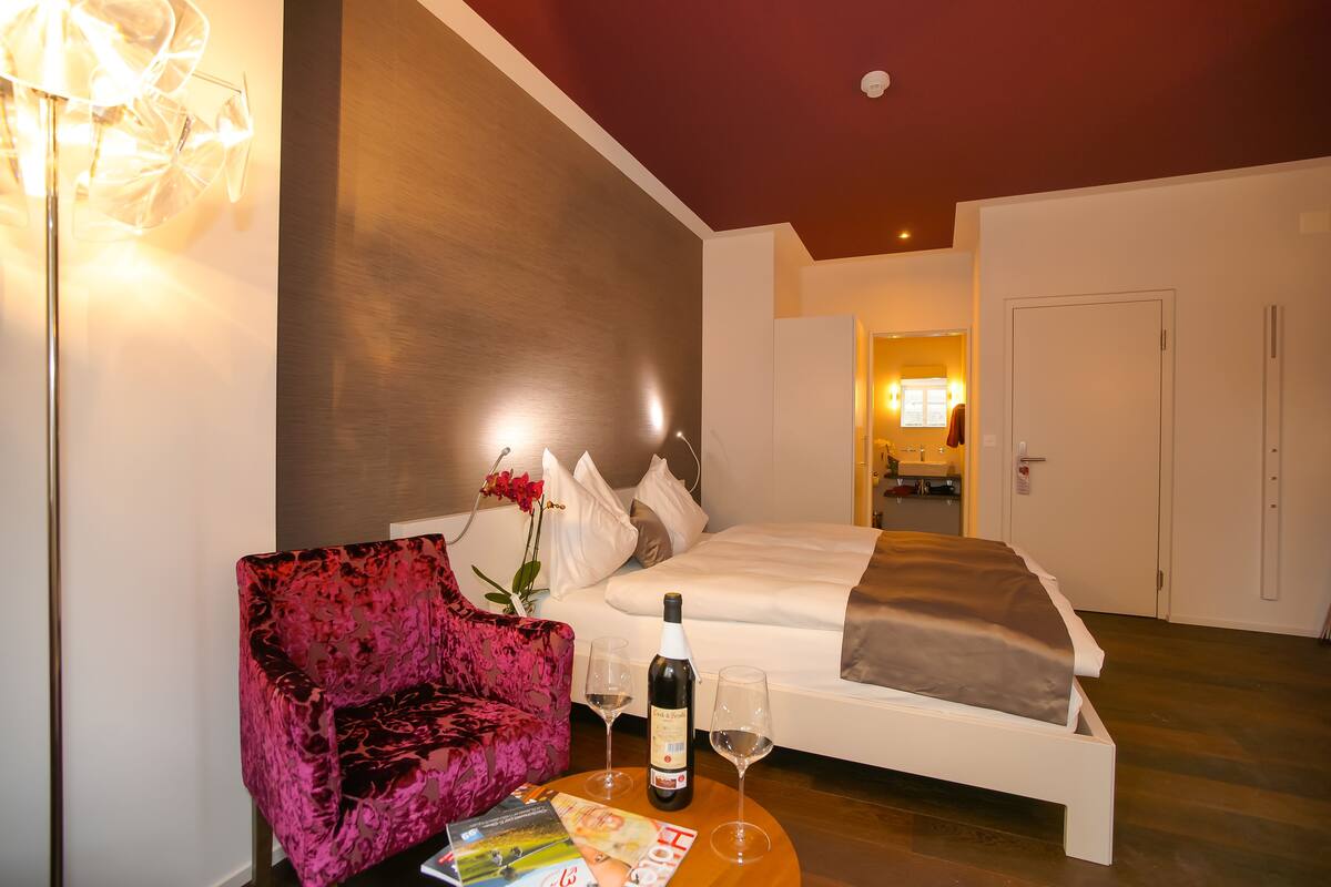 A hotel room with a bed, a pink chair, a table with a bottle and glasses, and a magazine.