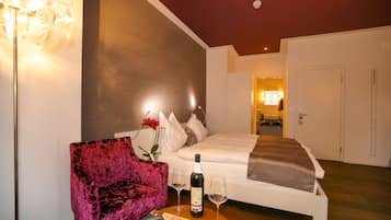 Quarto de hotel com uma cama, uma cadeira rosa, uma mesa com uma garrafa e copos, e uma revista.