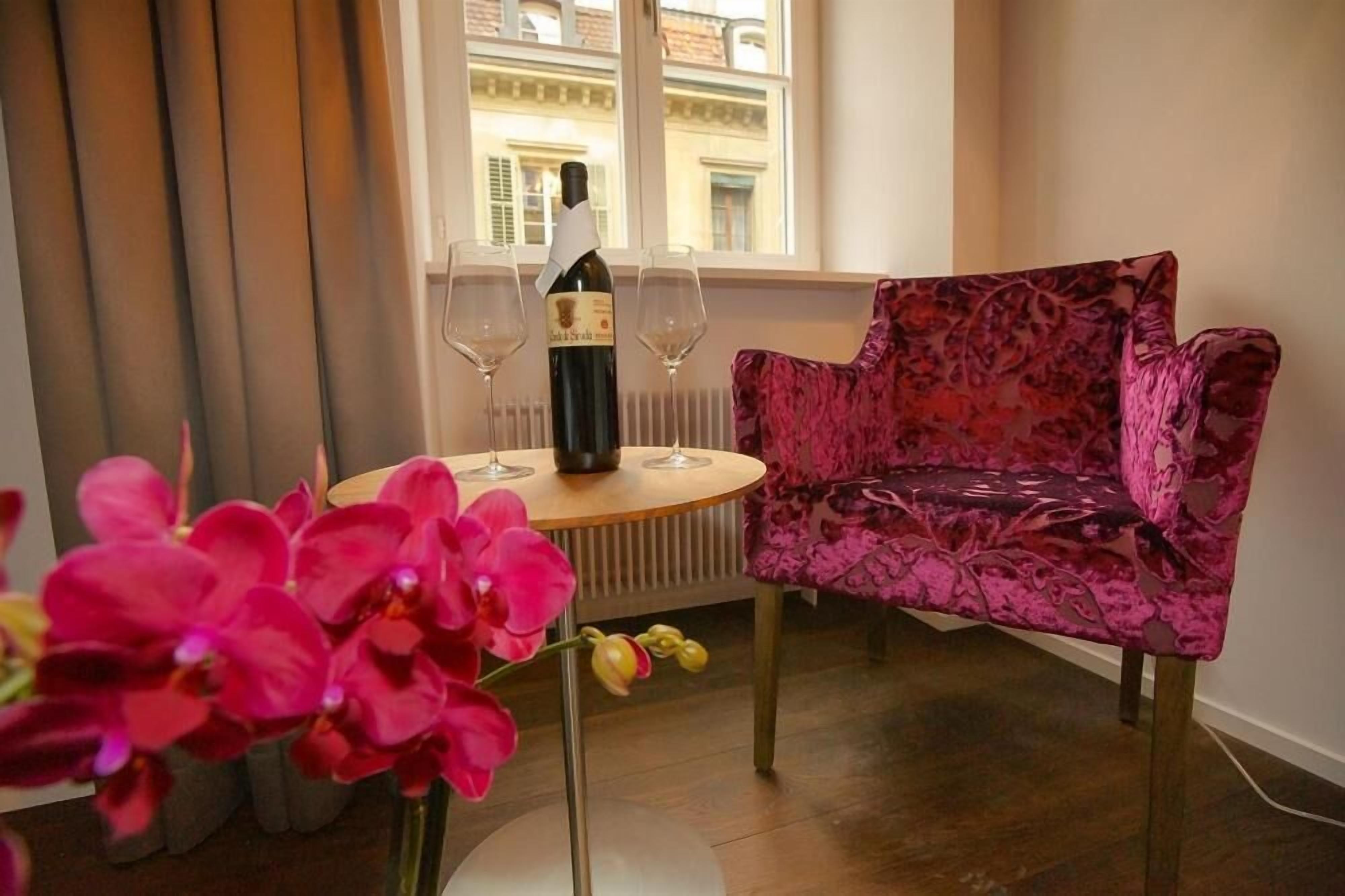 A room with a round table, a chair, a bottle of wine, and a window with a view of buildings.