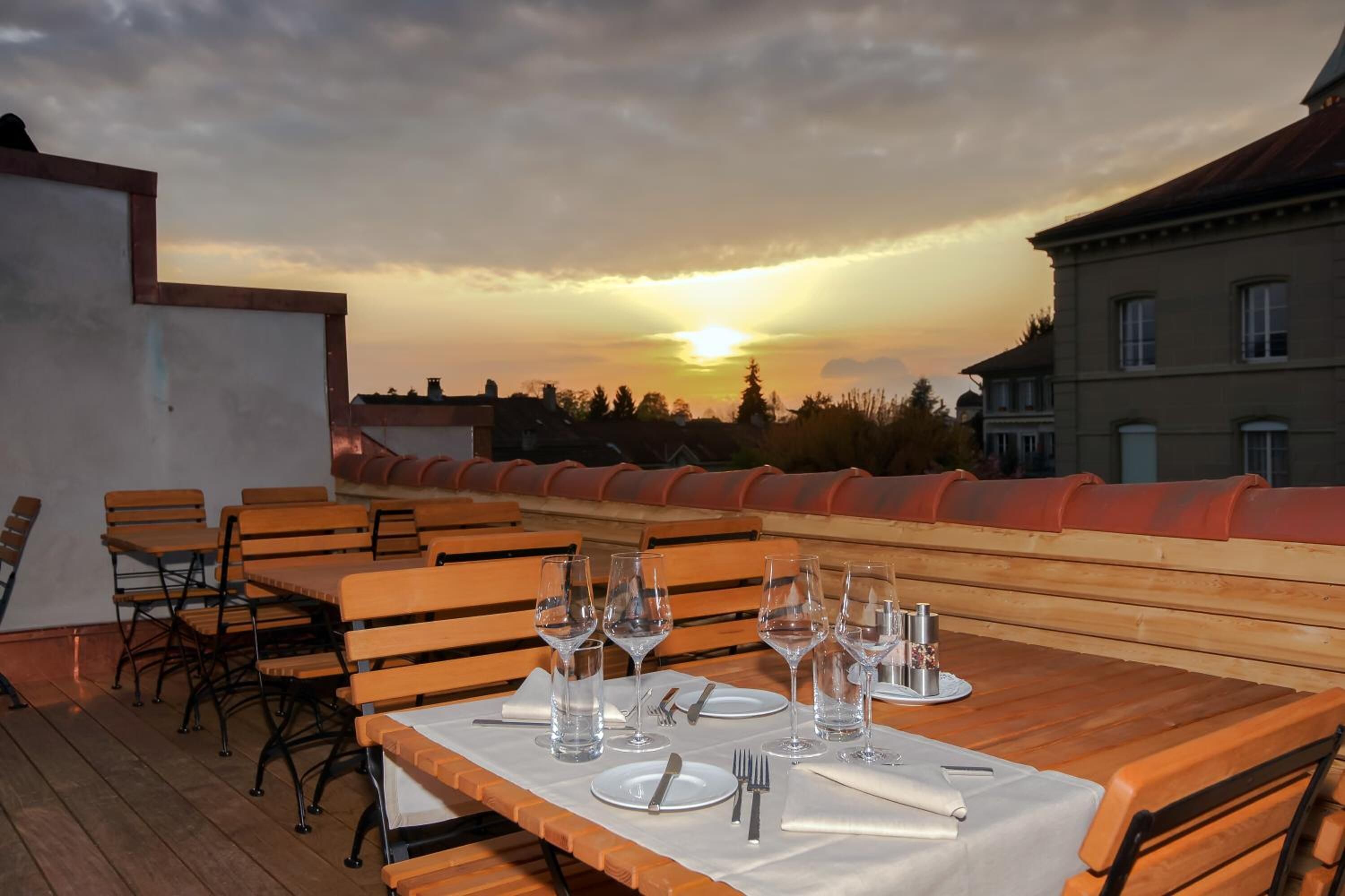 A rooftop dining area with tables set for four, each with wine glasses, water glasses, and cutlery. The setting features a sunset view over a cityscape.