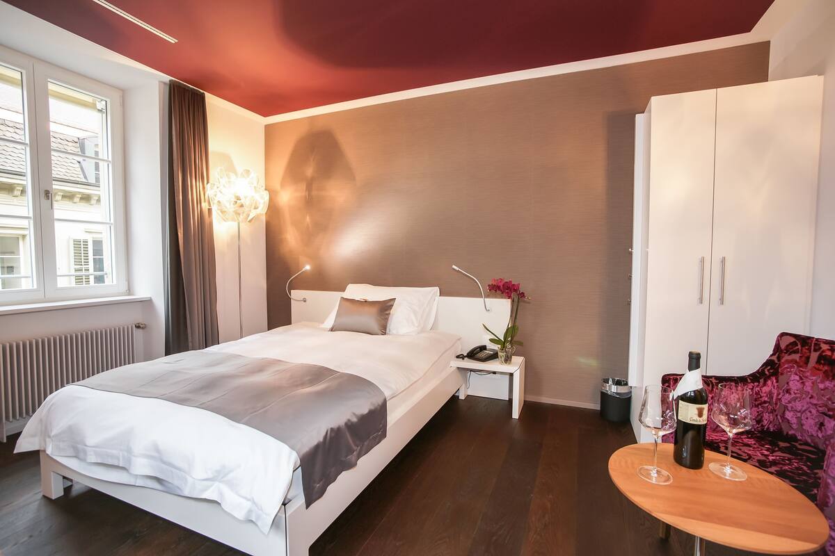 A hotel room with a bed, bedside table, a bottle of wine, two glasses, a nightstand, a window, a radiator, and a wardrobe.