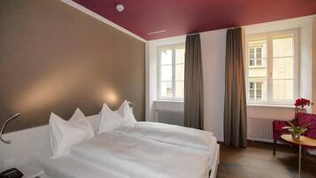 A hotel room with a bed, a bedside table with a lamp, a window with curtains, and a view of buildings outside.