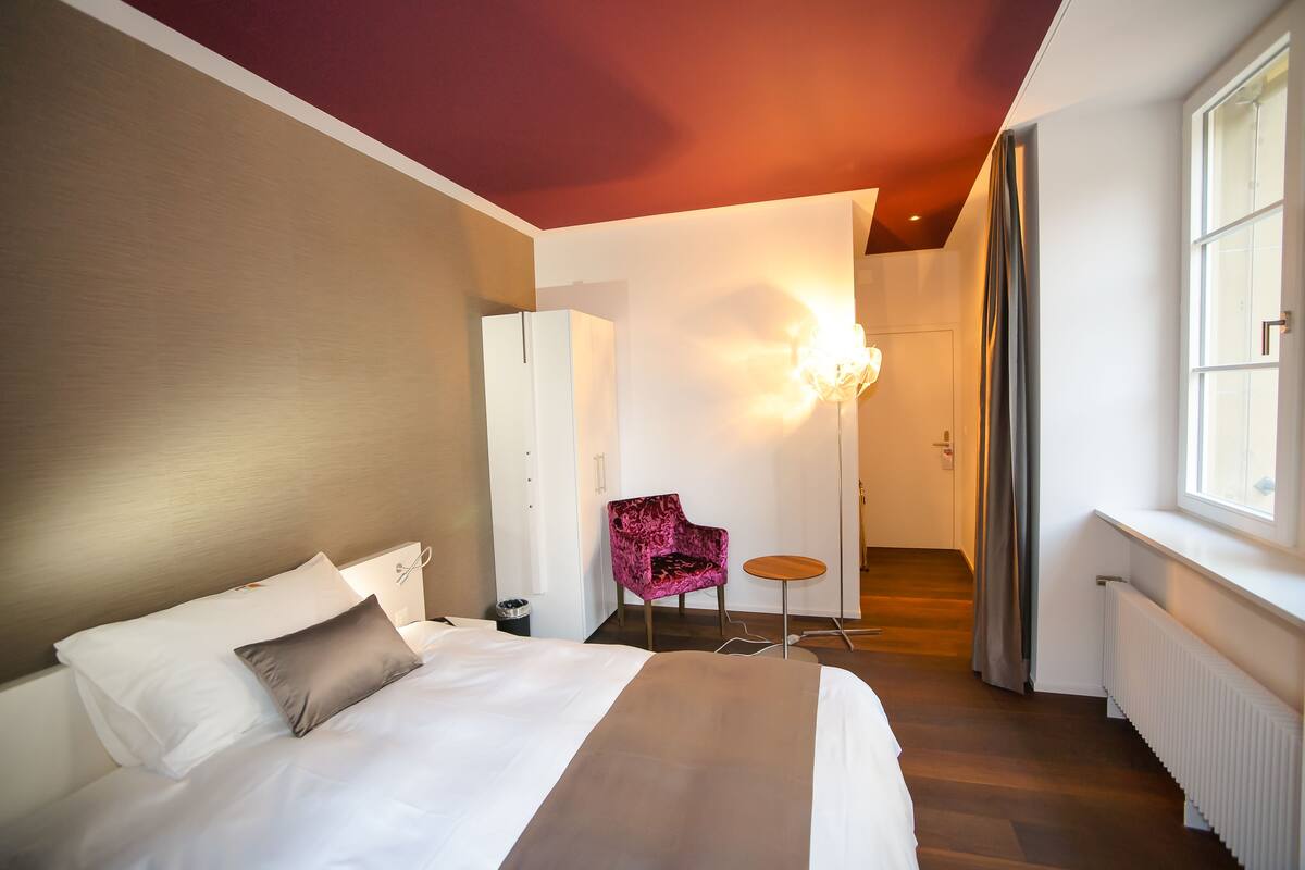 A hotel room with a bed, a chair, a small table, and a window.
