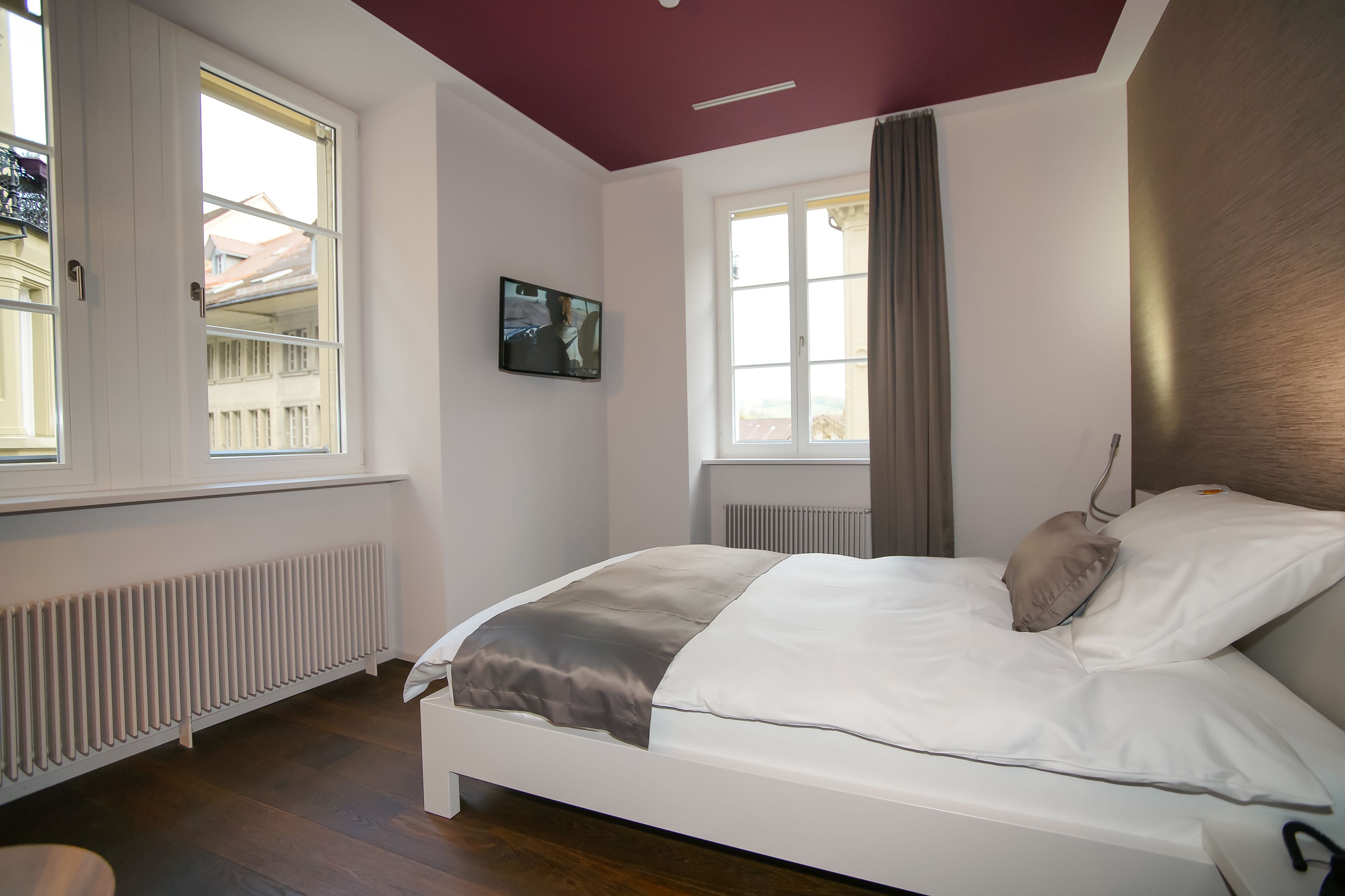 A bedroom with a bed, a TV mounted on the wall, and large windows with curtains.