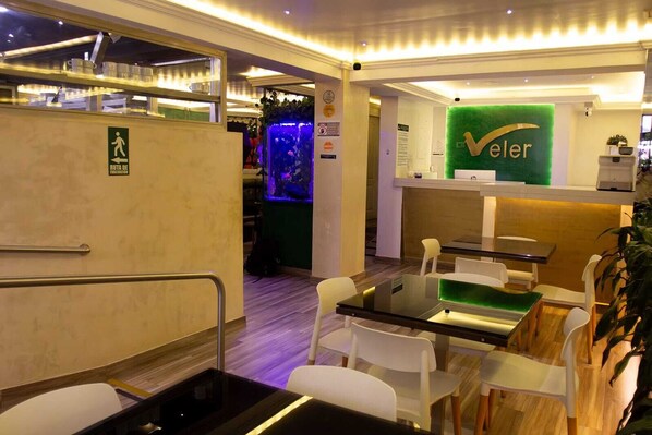 Vestíbulo de hotel con mostrador de recepción, pared verde con el logotipo 'Veler', separador de vidrio y señal de acceso para personas con discapacidad.