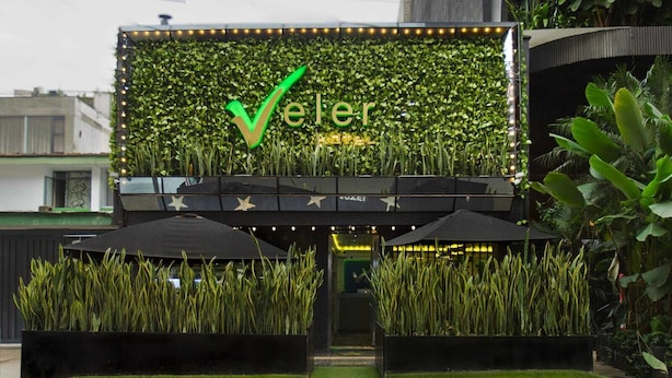 A building facade with a green leafy wall, a large checkmark logo, and two black umbrellas in front. - Hotel Veler (Medellín)