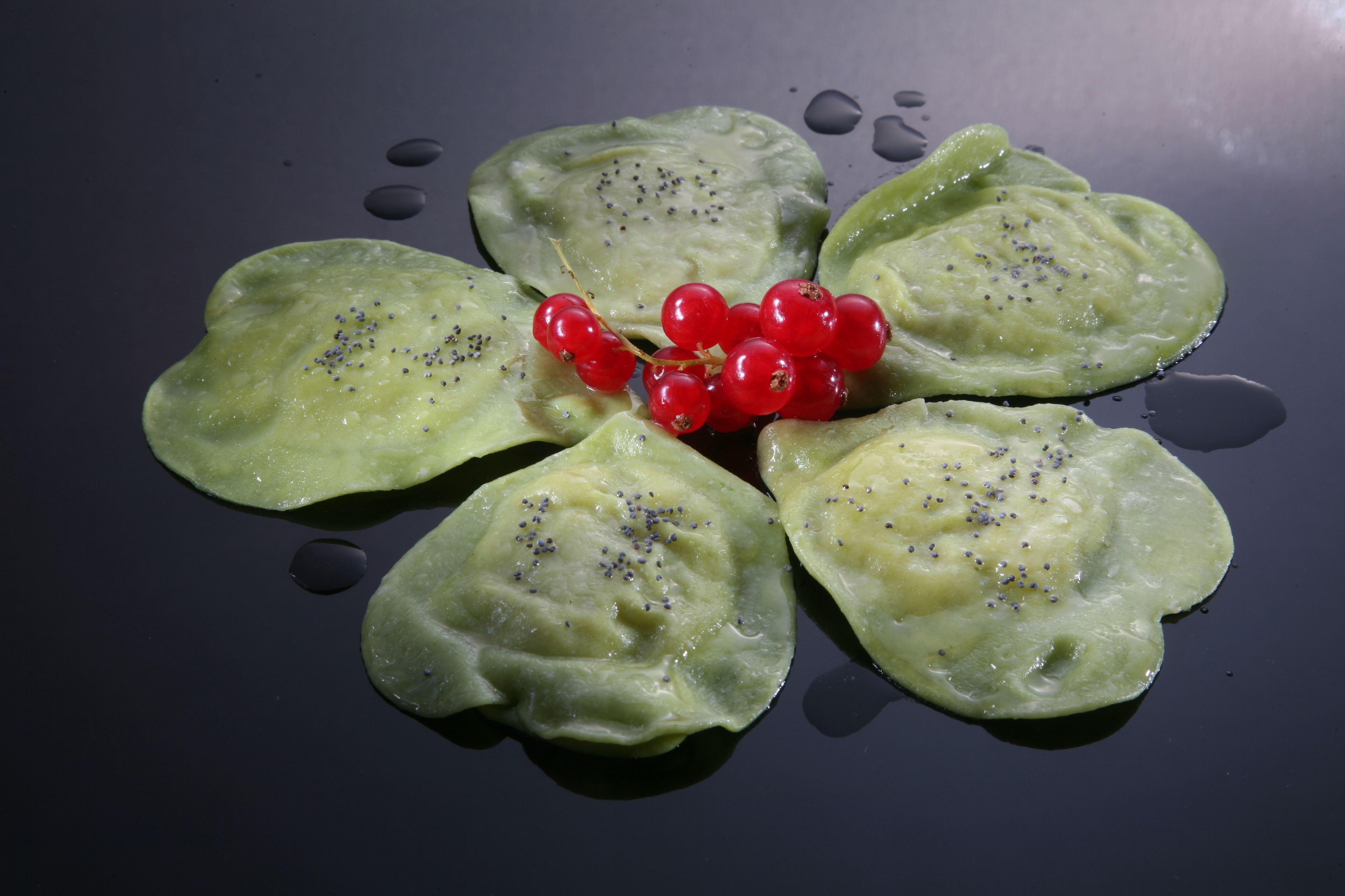Quattro ravioli con ripieno verde e macchie nere, disposti su una superficie scura con ribes rossi al centro.