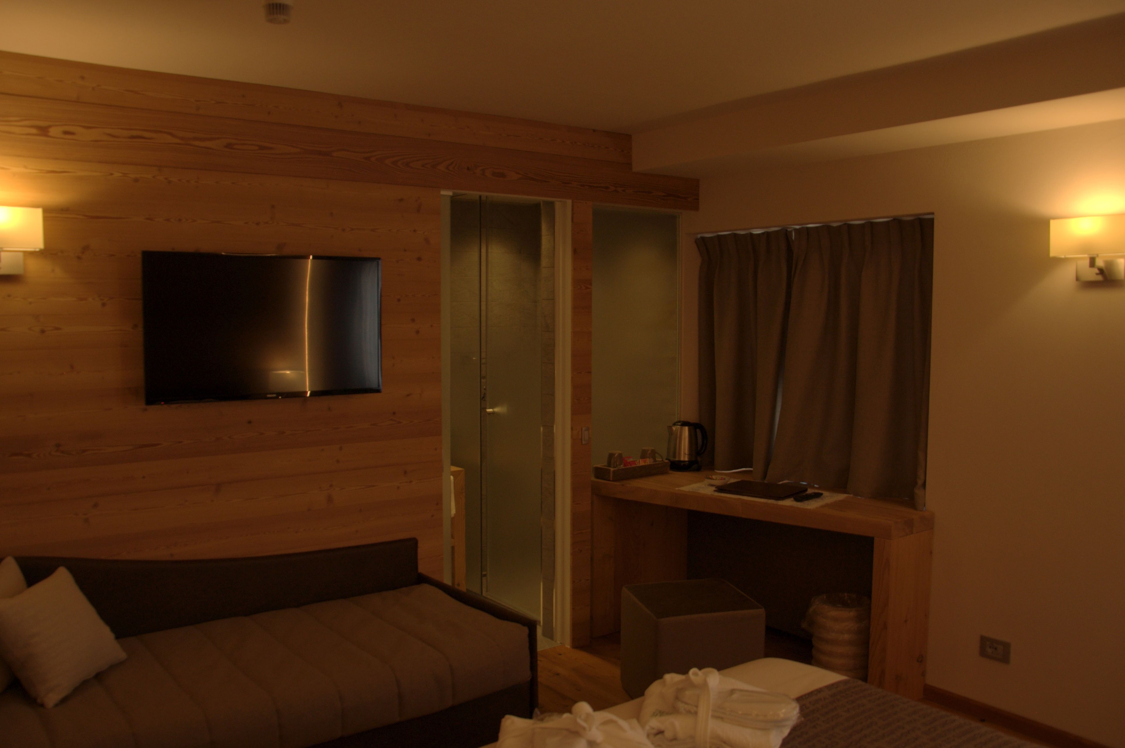 A room with a bed, a TV mounted on the wall, a small table with a kettle, and a window with curtains.