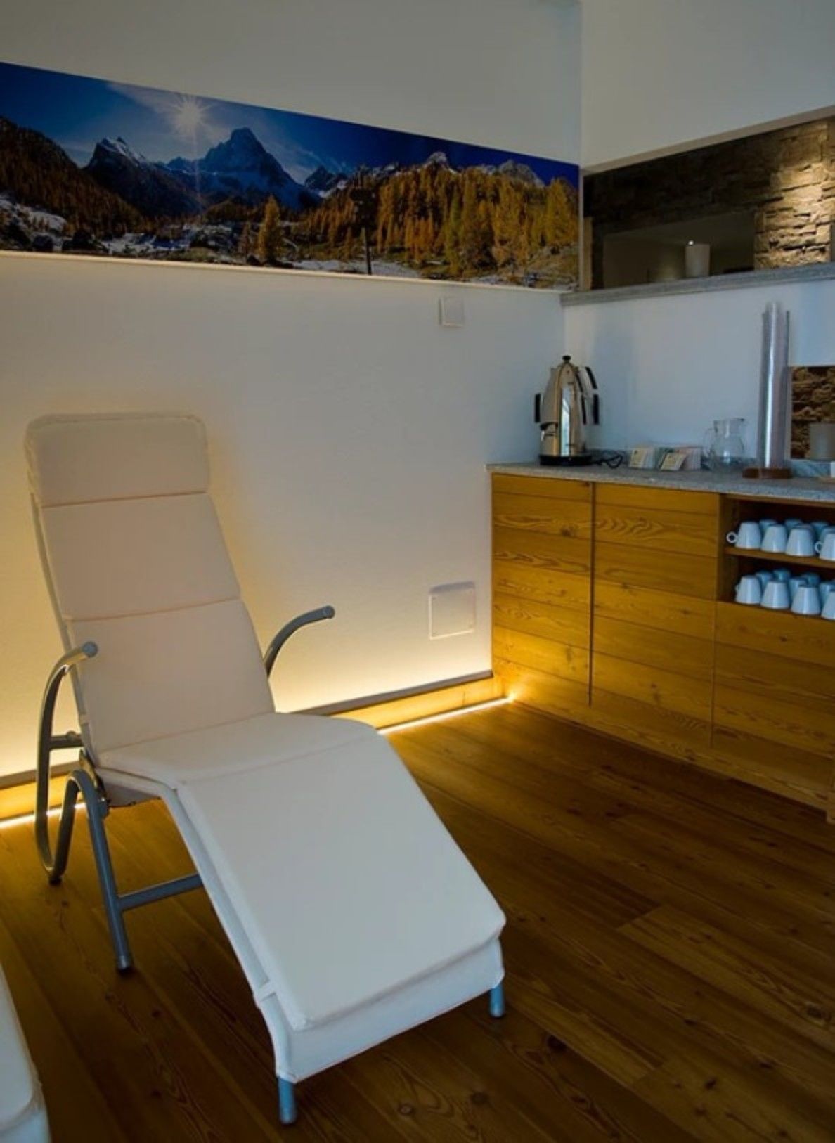A modern kitchen with wooden cabinets and a stone backsplash, a white chair with a backrest, and a mountain landscape mural on the wall.
