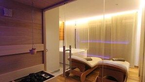 A modern spa room with a glass partition, wooden flooring, a massage table, a small shelf with a potted plant, and a massage chair. - Albergo Edelweiss (Crodo)