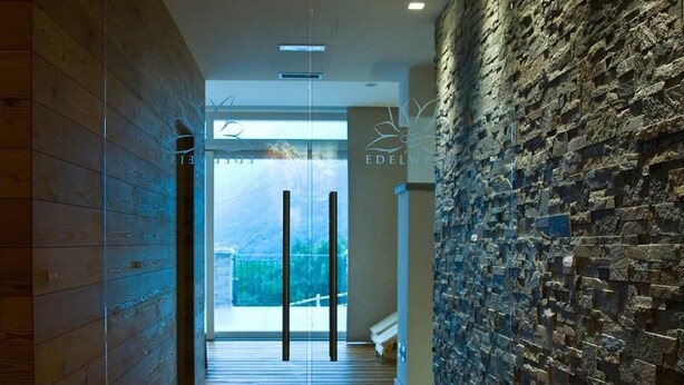 A modern hallway with wooden paneling on one side and a stone wall on the other, leading to a glass door with a view of greenery outside. - Albergo Edelweiss (Crodo)