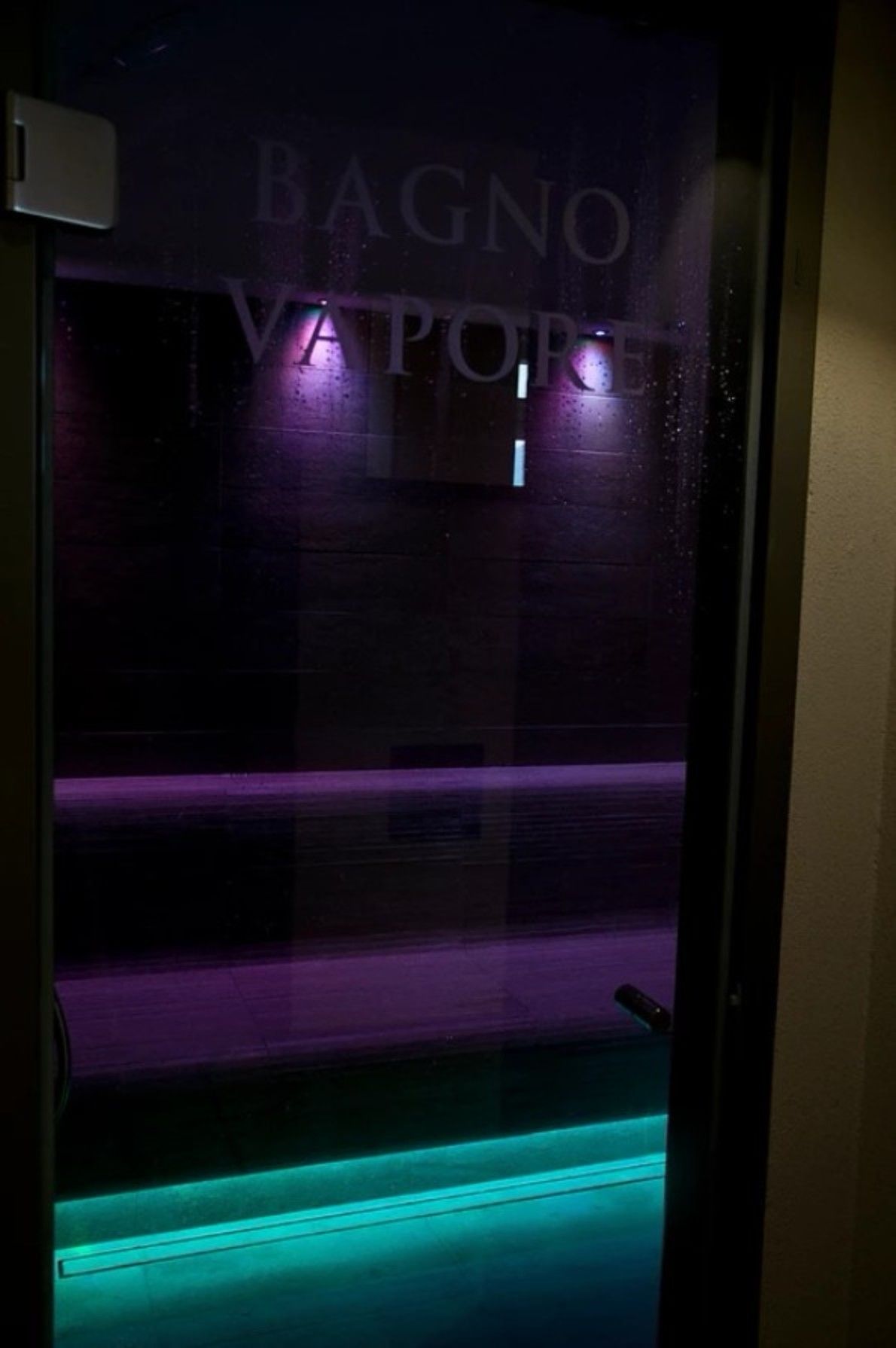Ascensor tenuemente iluminado con una superficie reflectante que muestra el texto 'BAGNO VAPOR' y una luz verde en la parte inferior.