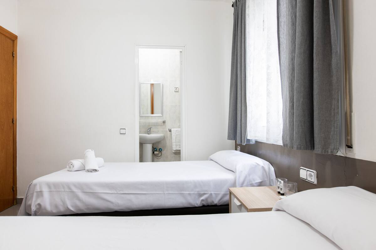 Een hotelkamer met twee bedden, een badkamer zichtbaar door een geopende deur en een raam met gordijnen.