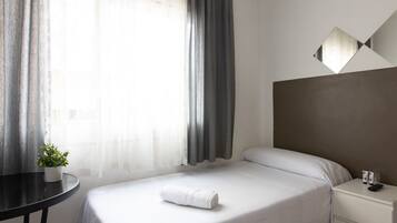 A hotel room with a bed, a bedside table with a plant, a nightstand with a remote control, and a wall-mounted light fixture.