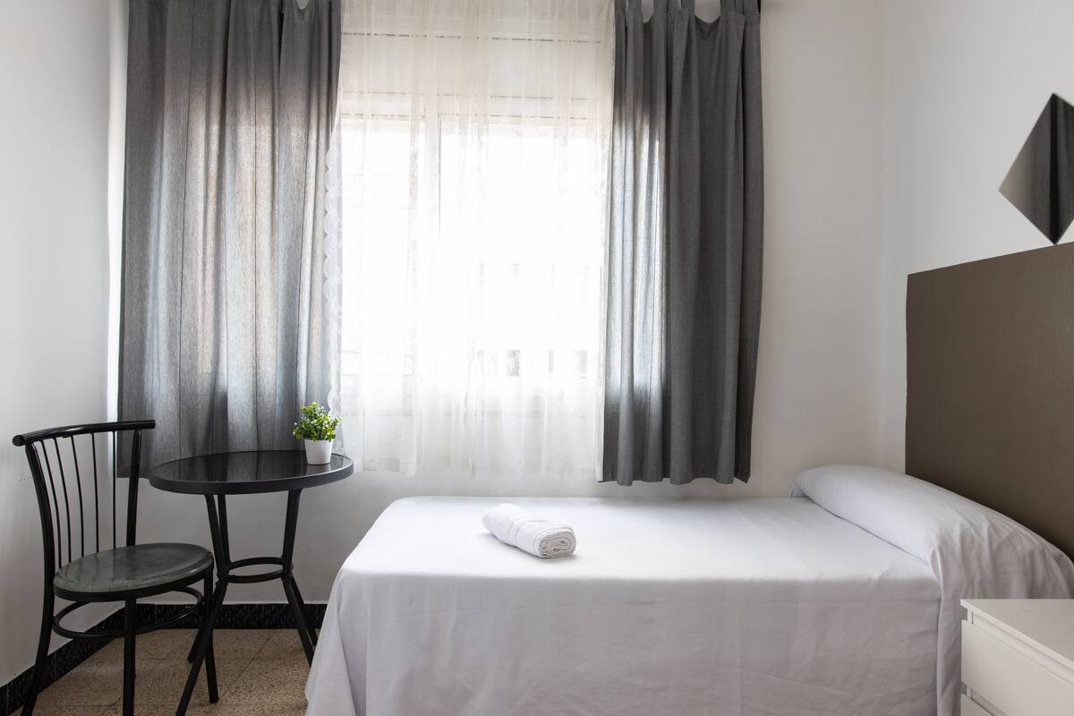 Een hotelkamer met een bed, een bijzettafeltje met een plant, een stoel en een raam met gordijnen.