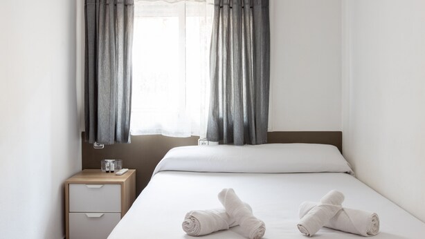 A hotel room with a bed, two rolled towels, a bedside table with a glass and a remote control, and grey curtains. - Pensión Galicia (Canovelles)