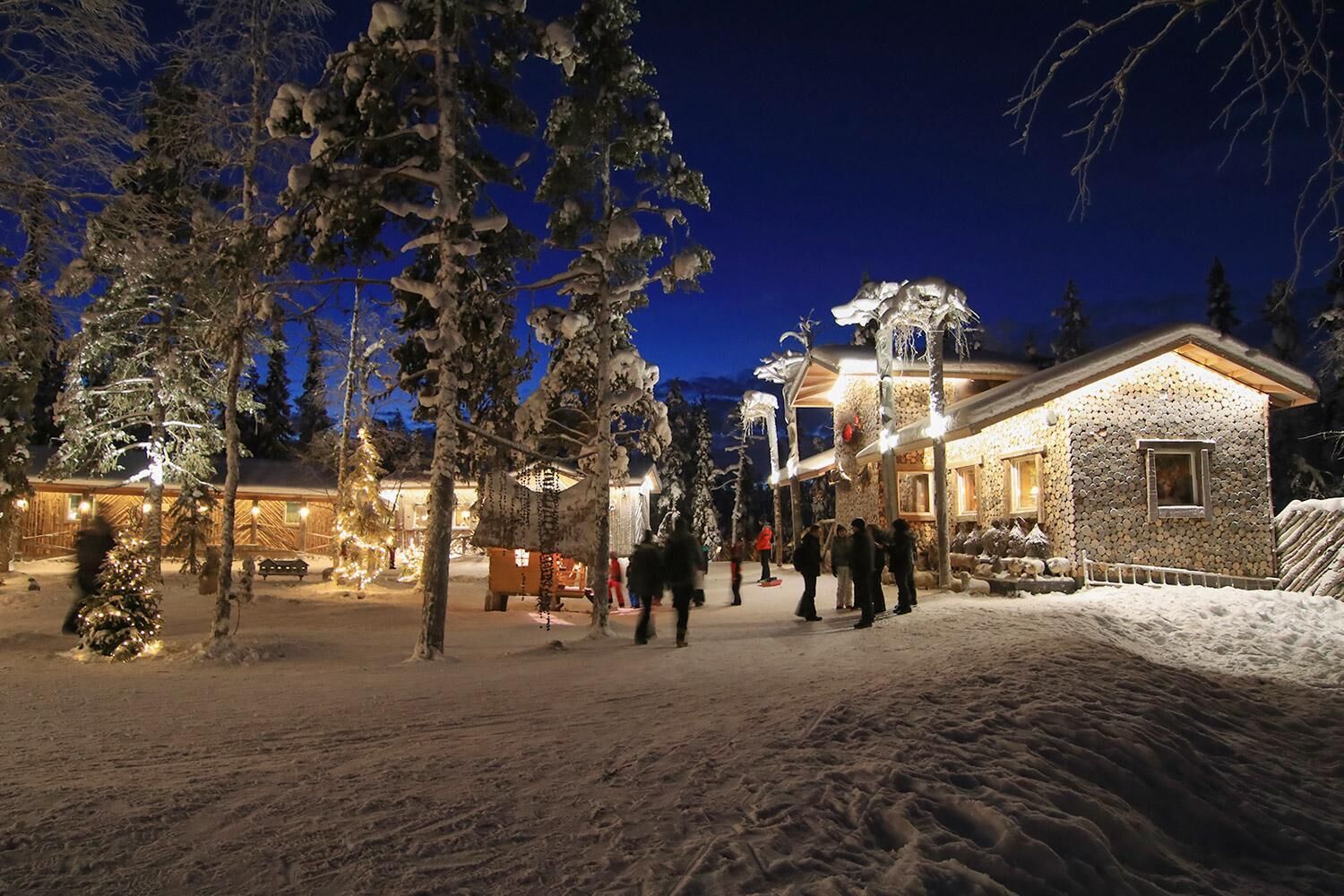 A snow-covered outdoor area with a building that has a stone facade and a wooden structure with lights.