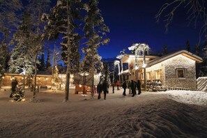 A snow-covered outdoor area with a building that has a stone facade and a wooden structure with lights. - Elves Village Hotel (Kittila)
