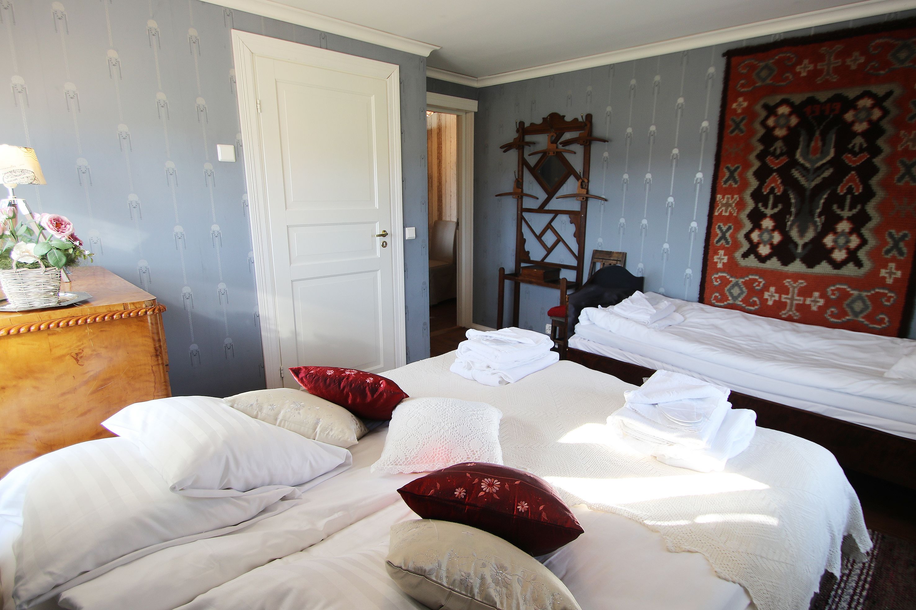 A neatly made bed with white and red pillows, a wooden dresser with a lamp and flowers, and a large patterned rug.