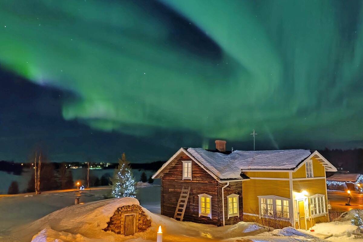 A wooden cabin with a snow-covered roof, a lit candle in the foreground, and a view of the Northern Lights in the background.
