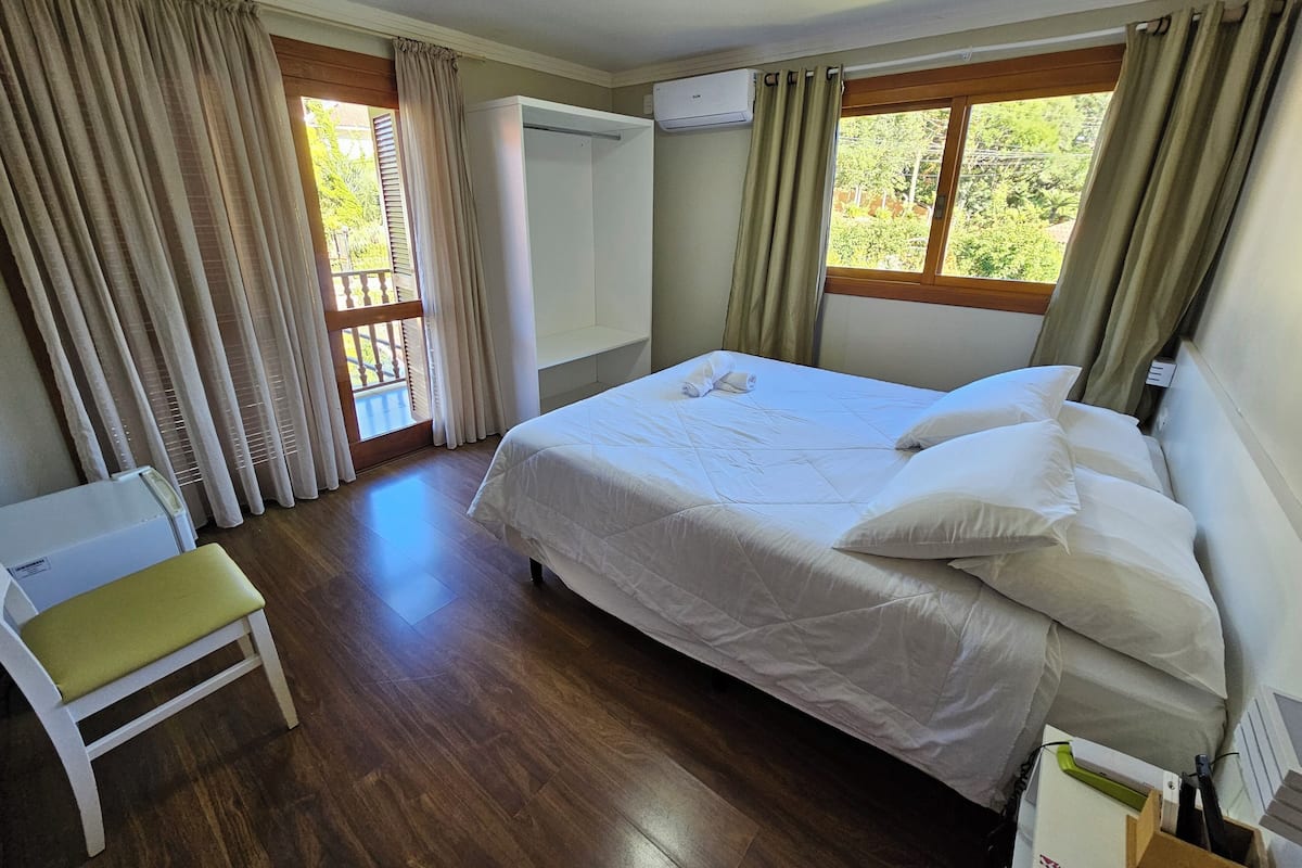 Un dormitorio con cama, una silla, ventana con cortinas y unidad de aire acondicionado.