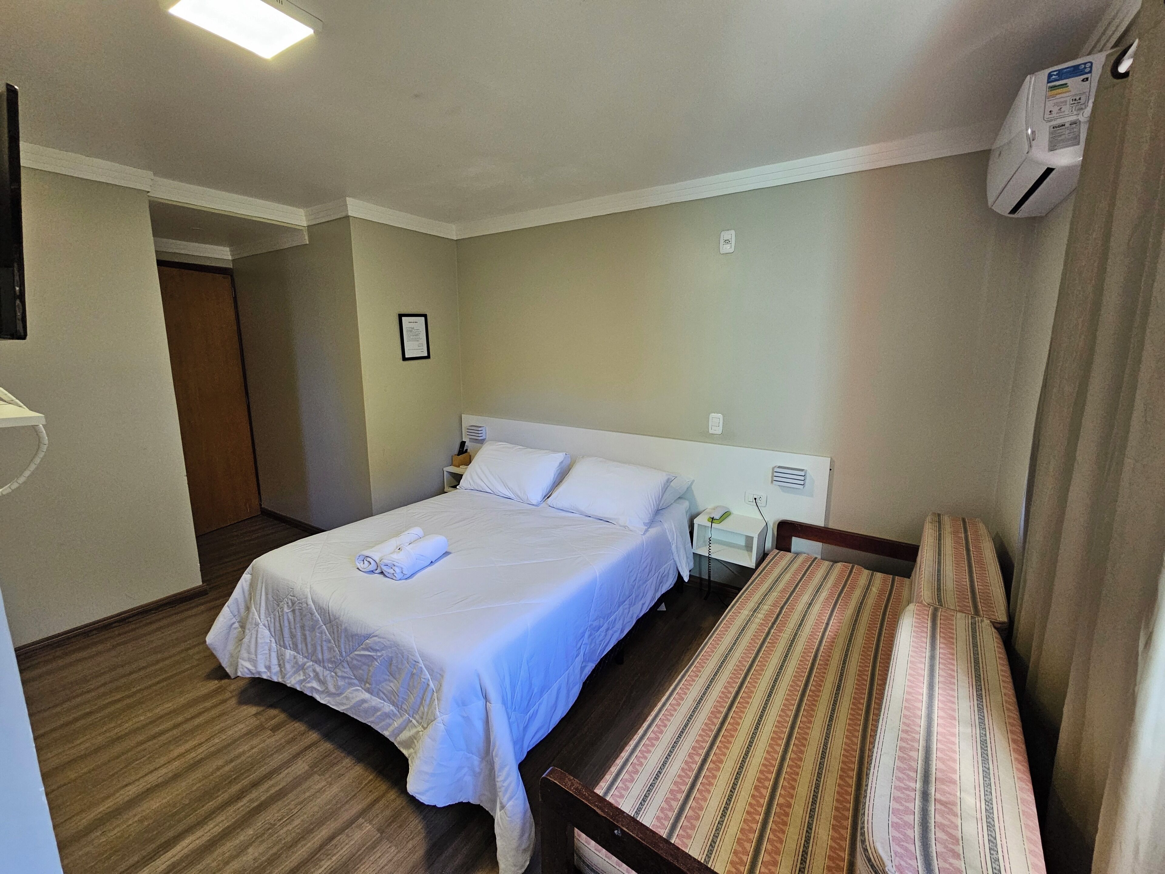 A hotel room with a bed, a bedside table, a lamp, a wall-mounted air conditioner, and a door.
