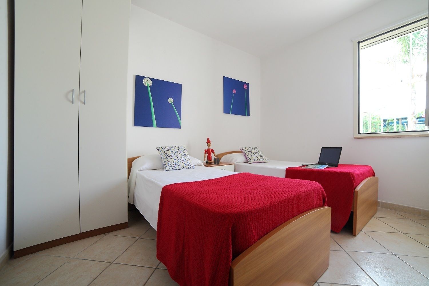 Een kamer met twee bedden, elk met een rode deken, wit beddengoed en een laptop op een van de bedden. Er zijn twee aan de muur bevestigde schilderijen, een houten garderobe en een raam met uitzicht.