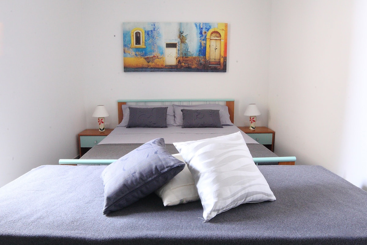 Una cama con ropa de cama y almohadas grises, un cuadro de un edificio en la pared y dos lámparas de noche.