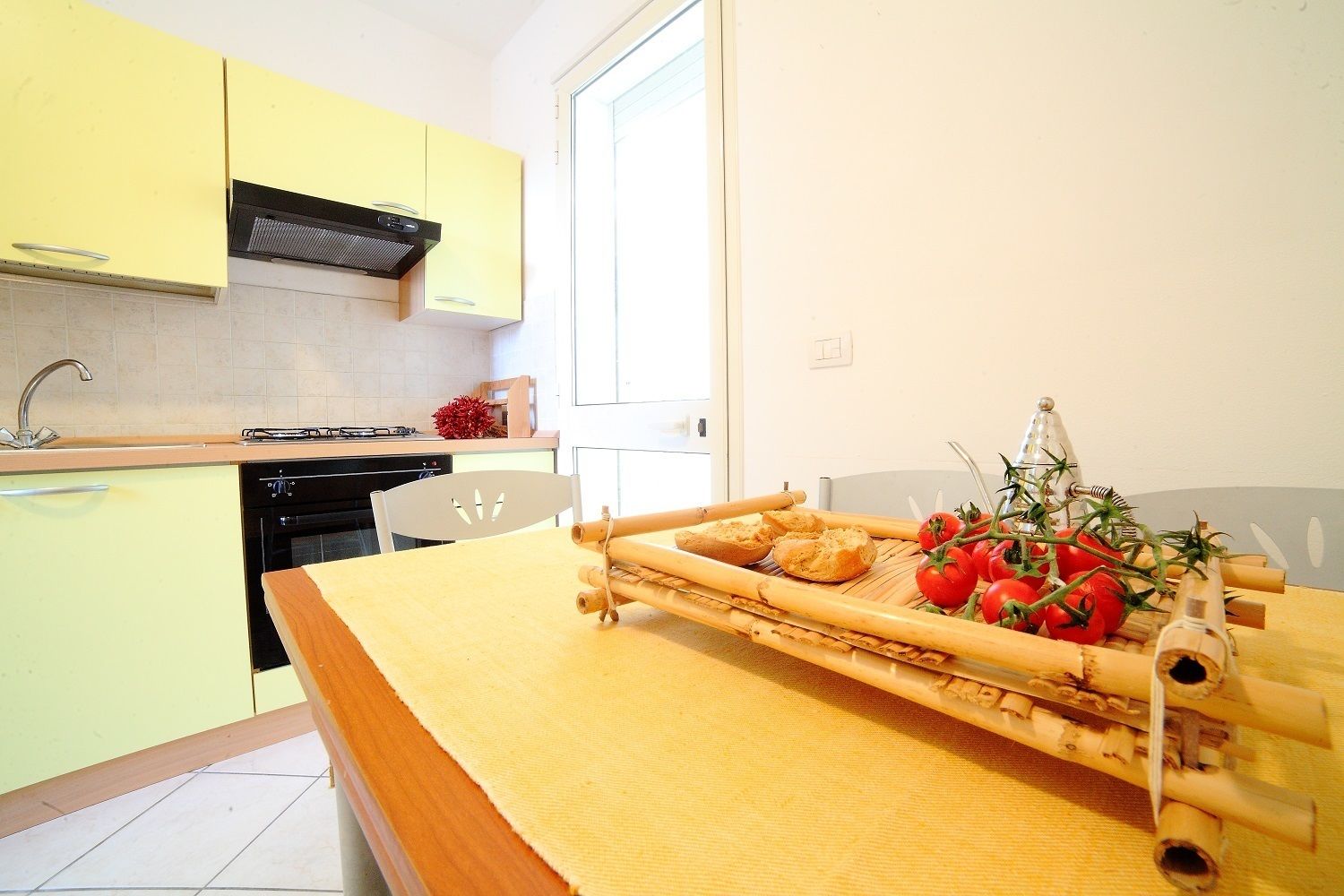 Una cocina con armarios amarillos, una estufa y una mesa con una bandeja de bambú que contiene pan y tomates.