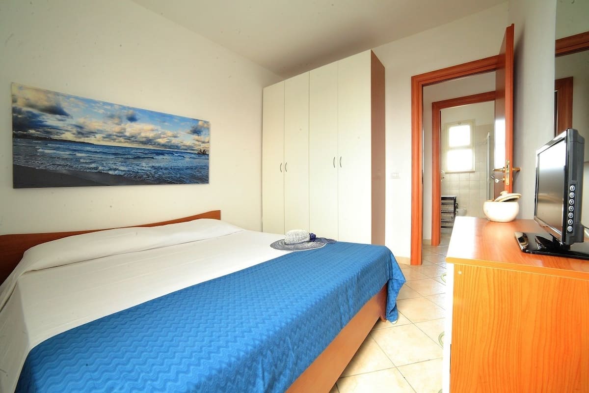 Habitación de hotel con cama, escritorio con televisor, armario y un cuadro con una escena de playa.
