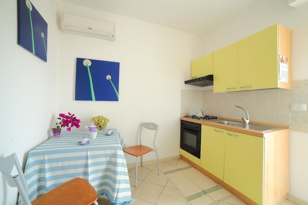 Una cocina compacta con armarios amarillos, una estufa a gas y un lavamanos. Hay una mesa de comedor con mantel azul y blanco y dos sillas. Un jarrón con flores está sobre la mesa.