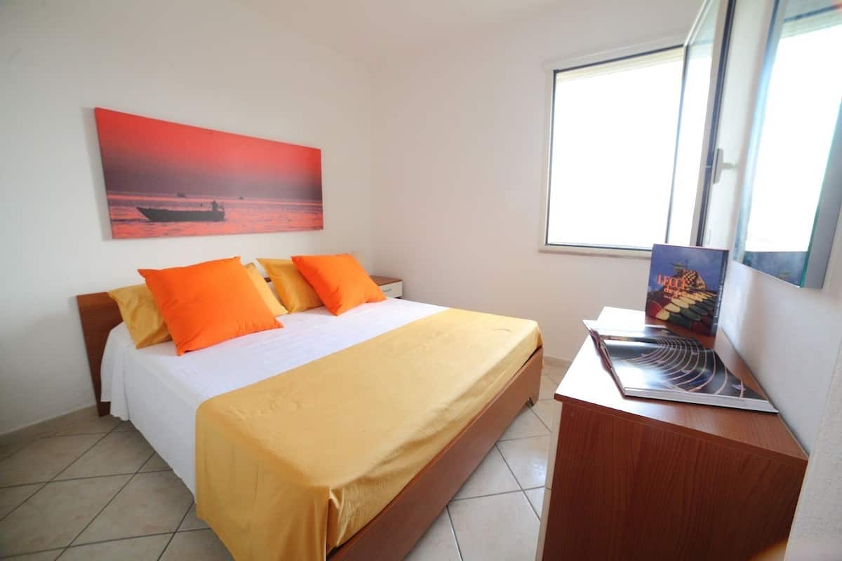 Un dormitorio con cama, mesita de noche, ventana y un cuadro en la pared.