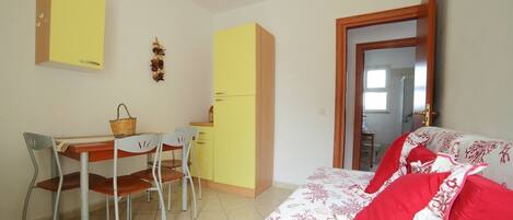 Una habitación pequeña y luminosa con una cama, un escritorio y sillas. Hay una ventana y una puerta.