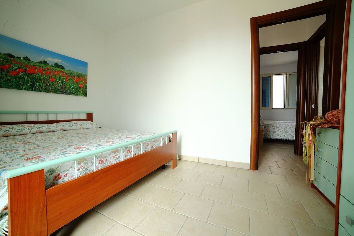 Un dormitorio con cama de madera, colcha estampada con flores, un cuadro de un campo y vistas a otra habitación a través de una puerta abierta.