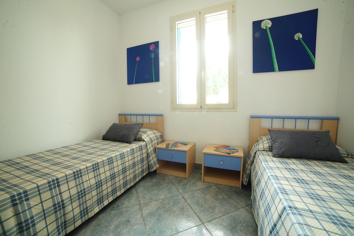 Una habitación pequeña y limpia con dos camas, cada una con cabecera azul, y una ventana que proporciona luz natural.