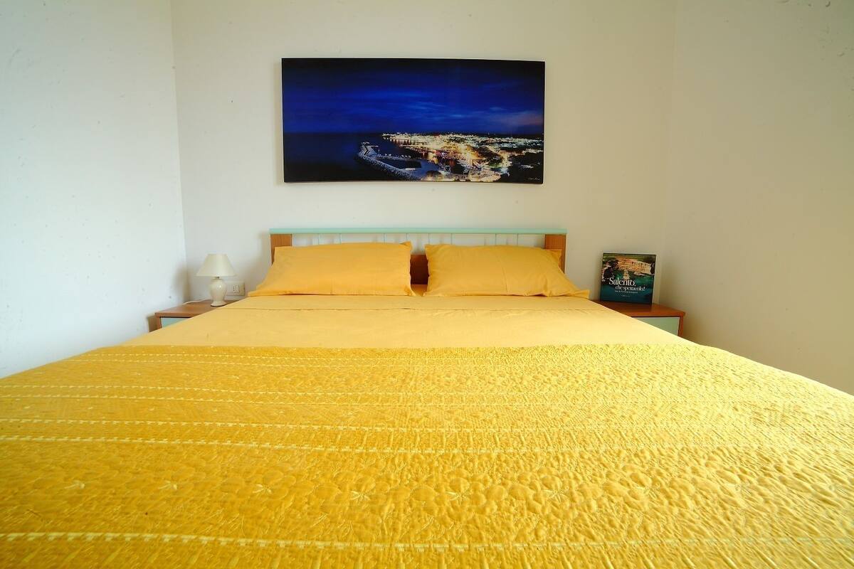 Una cama con colcha y almohadas amarillas, una mesita de noche con lámpara, y un cuadro en la pared con una vista urbana.
