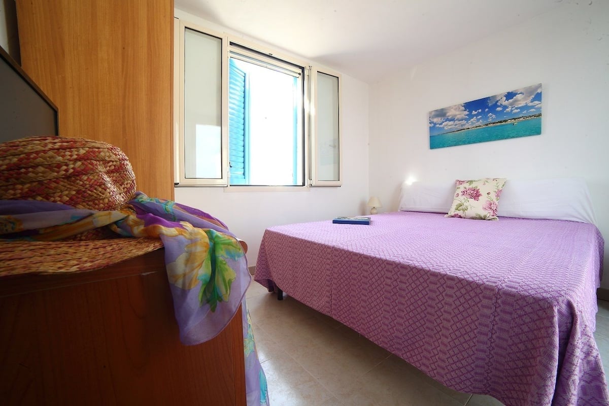 Un dormitorio con cama, una ventana con persianas, un cuadro de una playa y una cesta tejida sobre un aparador de madera.