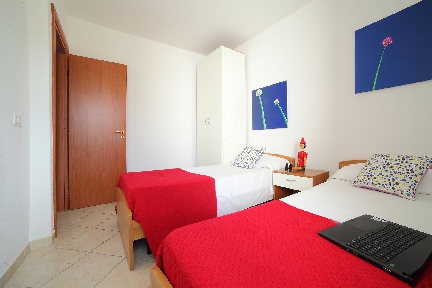 A bedroom with two beds, a wooden door, a nightstand with a lamp, and a laptop on one bed.