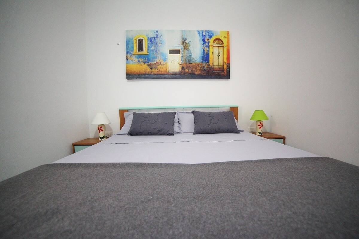 Una cama bien hecha con un edredón gris, dos lámparas de noche y un cuadro de un edificio en la pared.