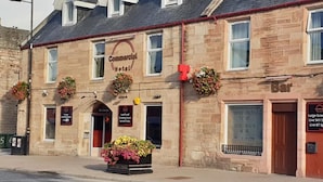 A stone building with a sign reading 'Commercial Hotel.' There are flower baskets on the windowsills and a flower planter in front. - COMMERCIAL HOTEL (Alness)