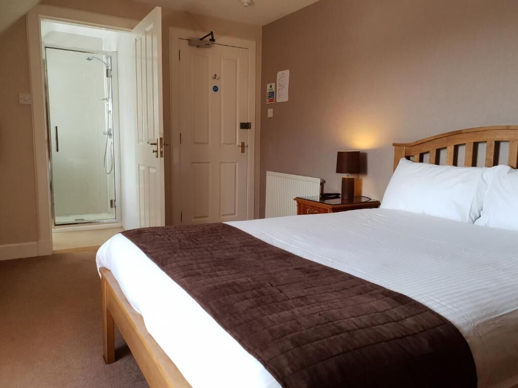 Habitación de hotel con cama, mesita de noche con lámpara, puerta que da a una ducha y un radiador.