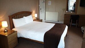 A neatly made bed with a white and brown blanket, two bedside lamps, a wooden nightstand, a wooden dresser, a radiator, and a mirror on the wall. - COMMERCIAL HOTEL (Alness)