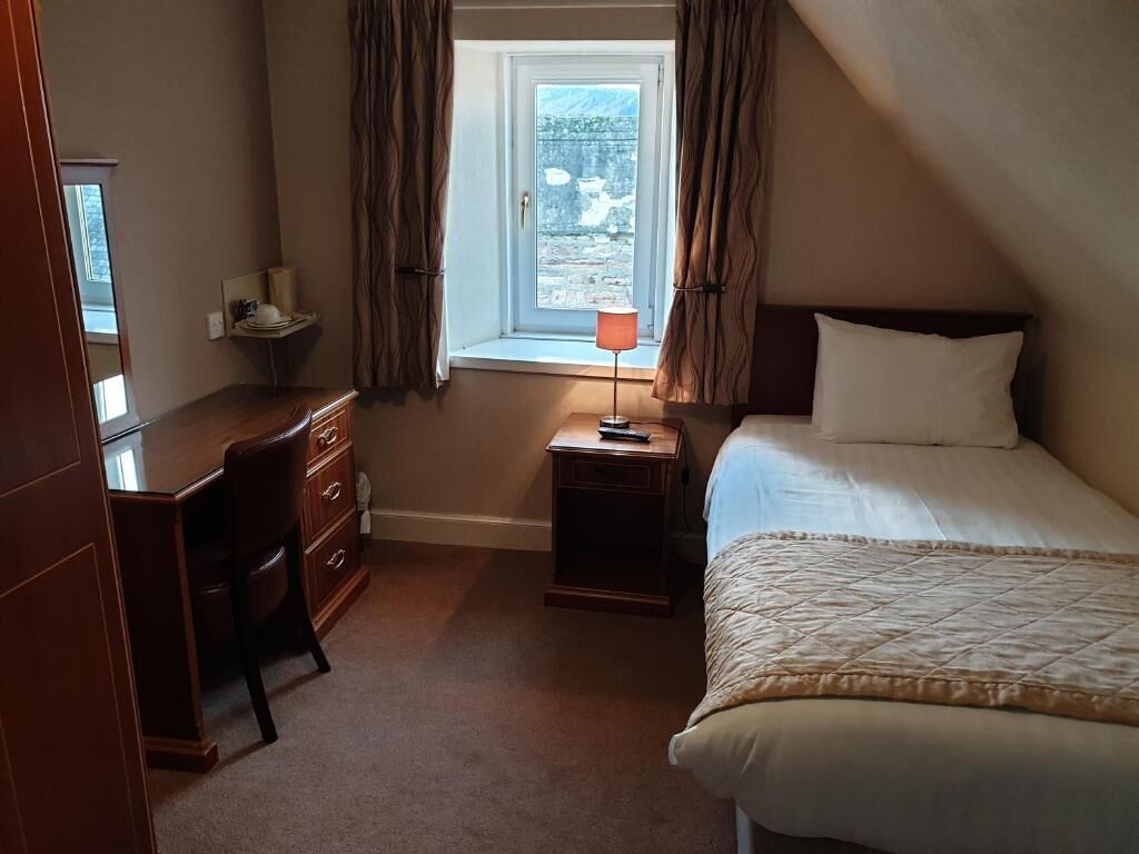 A bedroom with a bed, a desk, a chair, a nightstand with a lamp, and a window with curtains.