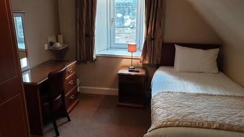 A bedroom with a bed, a desk, a chair, a nightstand with a lamp, and a window with curtains.