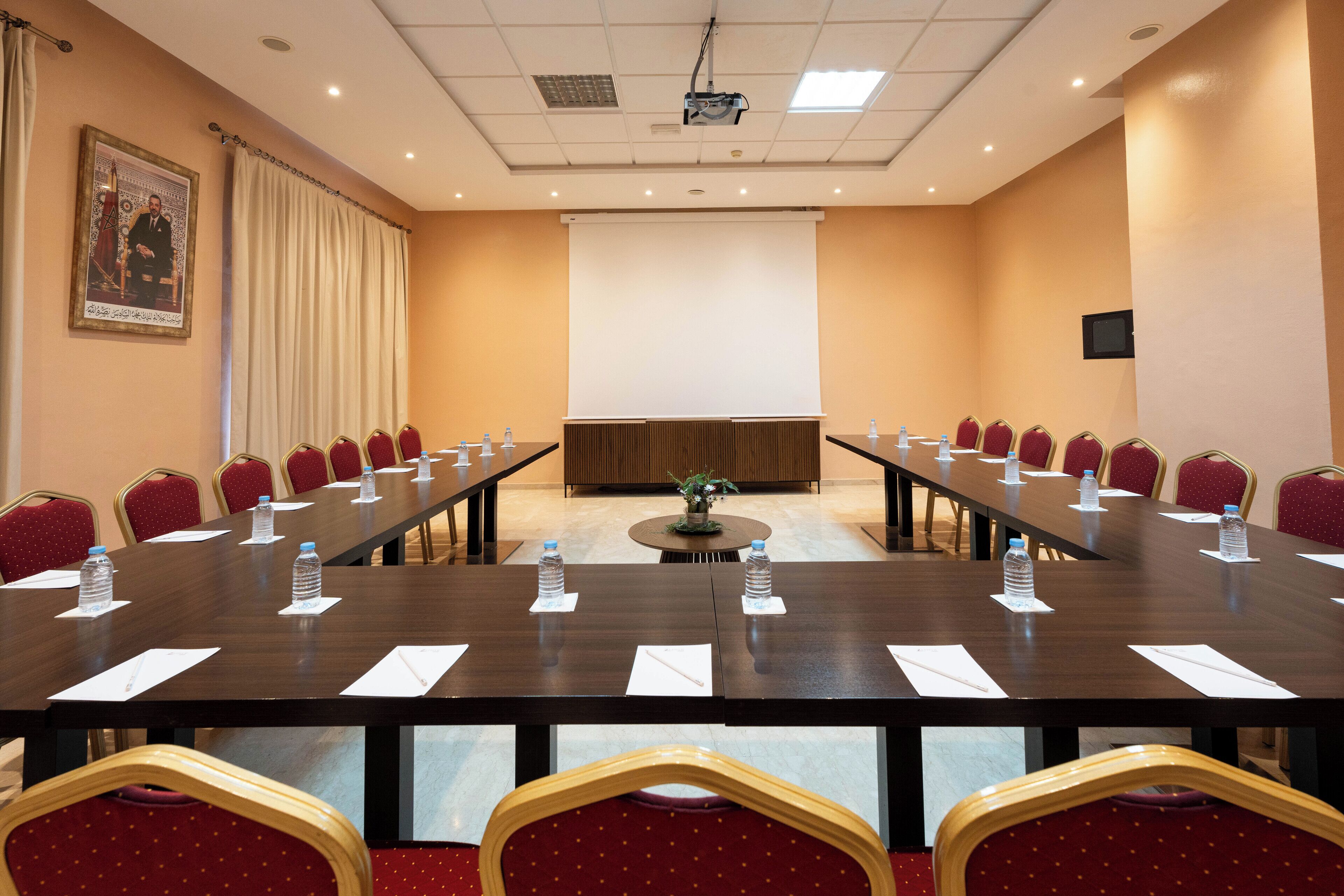 Un salón de conferencias con una larga mesa, sillas rojas, botellas de agua y una pantalla para proyector.