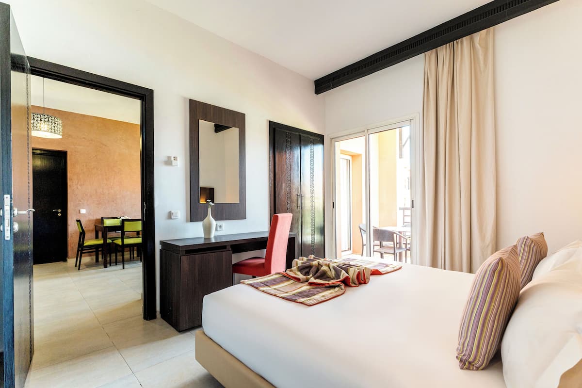 A hotel room with a large bed, a desk, and a chair. There is a door leading to a dining area.